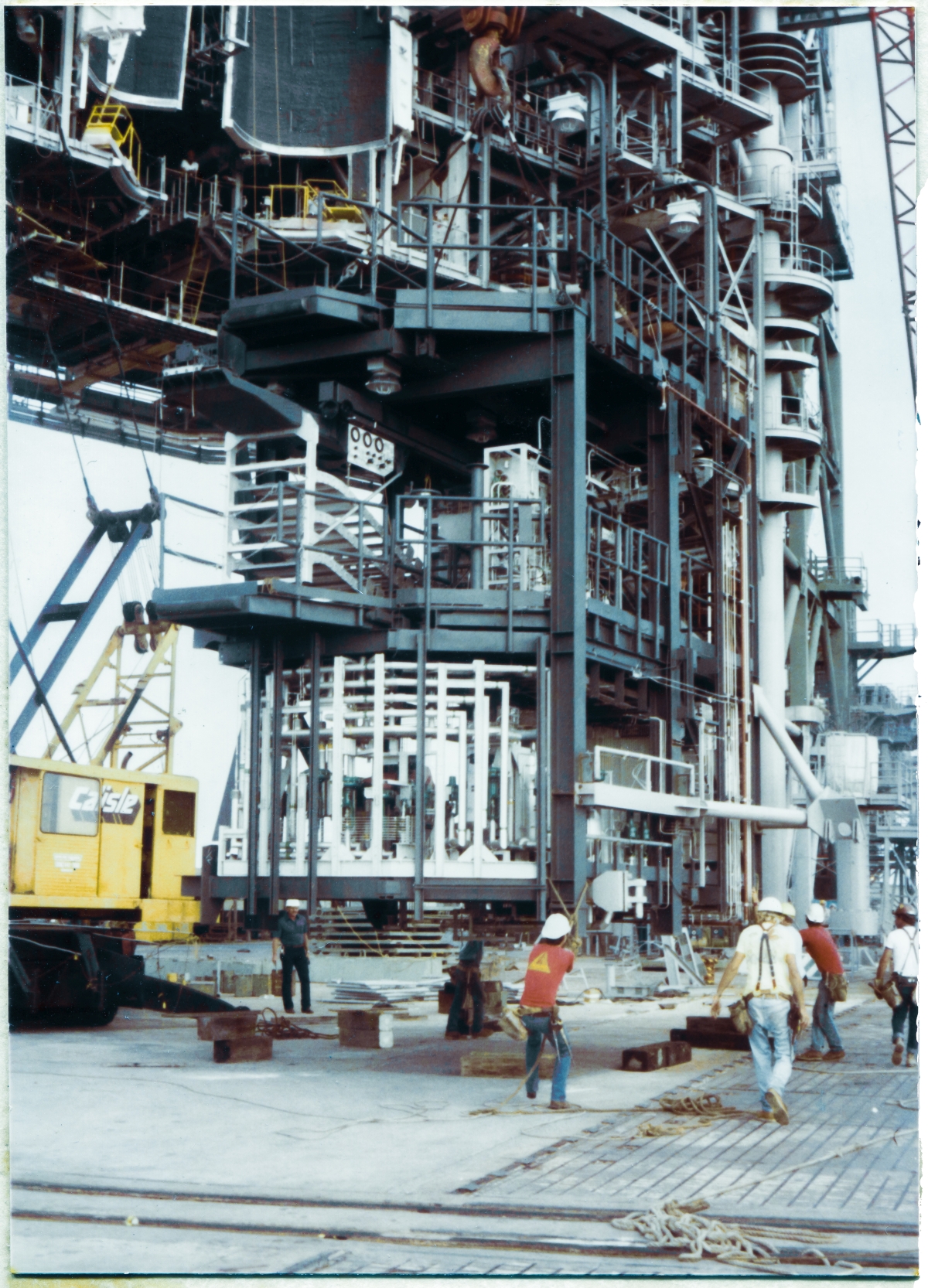 Image 088. Union Ironworkers from Local 808, working for Ivey Steel at Space Shuttle Launch Complex 39-B, Kennedy Space Center, Florida, are hands-on with the OMBUU after it was handed off from the smaller crane which took it from its original position on the Pad Deck near Column Line 7 under the RSS, and connected to the big Manitowoc which is going to take it up to its final elevation, well over a hundred feet above where you see it in this photograph. The crane operator continues to work it northward across the Pad Deck, but very soon it will begin moving vertically, once they get it to the place directly beneath where it needs to be lifted straight up from. Once it has been lifted to its final elevation, things will be very carefully looked over, making sure all clearances and fits are properly maintained and aligned, and then it will be very gently swung in toward the face of the RSS, where the ironworkers, working with the crane operator, will fine-position it and bolt it into place against the connection plates it will be attached to. Photo by James MacLaren.